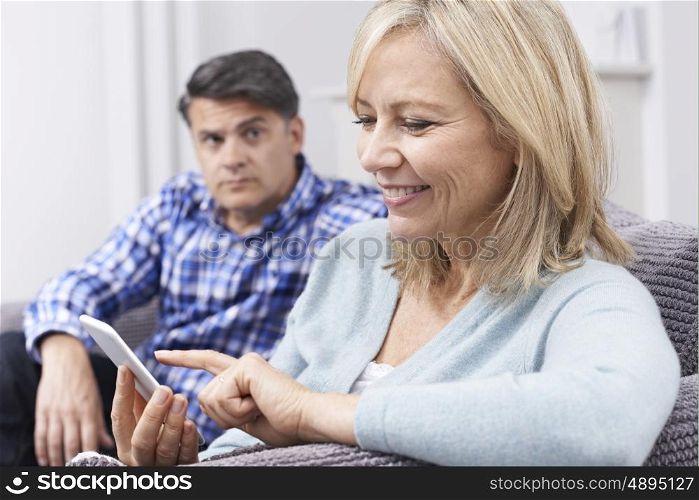 Unhappy Man Sitting On Sofa As Partner Uses Mobile Phone
