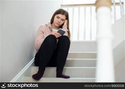 Unhappy Teenage Girl Being Bullied By Text Message Sitting On Stairs At Home