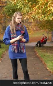 Unhappy Teenage Girl Standing In Autumn Park With Couple On Bench In Background