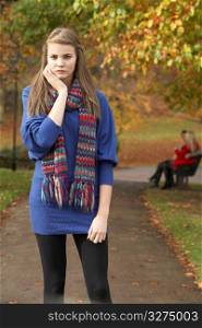 Unhappy Teenage Girl Standing In Autumn Park With Couple On Bench In Background