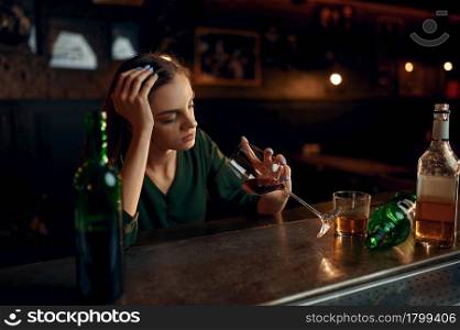 Unhappy woman drinks alcohol beverage at the counter in bar. One female person in pub, human emotions, leisure activities, depression. Unhappy woman drinks alcohol at the counter in bar