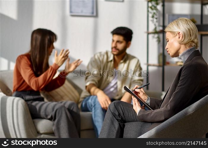 Unhappy young couple arguing at psychologists office. Focus on therapist making notes in clipboard. Unhappy young couple arguing at psychologists office