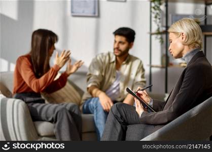 Unhappy young couple arguing at psychologists office. Focus on therapist making notes in clipboard. Unhappy young couple arguing at psychologists office