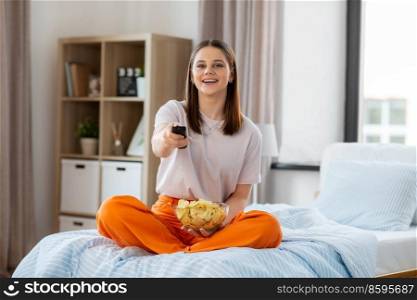 unhealthy eating, fast food and people concept - happy girl with remote control and crisps watching tv sitting on bed at home. happy girl with crisps watching tv at home
