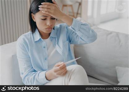 Unhealthy female holding thermometer, checking body temperature, suffers from flu or coronavirus at home. Sick woman has covid symptom, touches hot forehead, suffering from fever, frowning.. Unhealthy female holding thermometer, checking body temperature, suffers from flu or coronavirus 