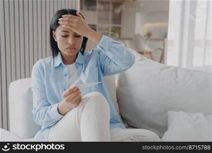Unhealthy female holds electronic thermometer suffering from flu or coronavirus fever, high body temperature. Ill young woman has covid symptom, touching hot forehead, sitting on sofa.. Unhealthy female holds thermometer, suffering from flu or coronavirus fever, high body temperature