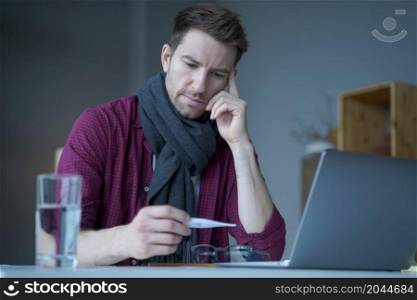 Unhealthy looking male freelancer feeling unwell and cold, wearing warm knitted scarf around neck, looking on digital thermometer in his hand checking temperature level while working remotely online. Unhealthy looking male freelancer feeling unwell and cold, wearing warm knitted scarf around neck