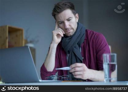 Unhealthy looking male freelancer feeling unwell and cold, wearing warm knitted scarf around neck, looking on digital thermometer in his hand checking temperature level while working remotely online. Unhealthy looking male freelancer feeling unwell and cold, wearing warm knitted scarf around neck