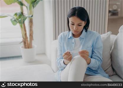 Unhealthy sad woman suffer from high body temperature, fever, holding thermometer. Sick worried young female suffering from flu, cold or coronavirus sitting on sofa at home.. Unhealthy young woman suffer from high body temperature, holding thermometer sitting on sofa