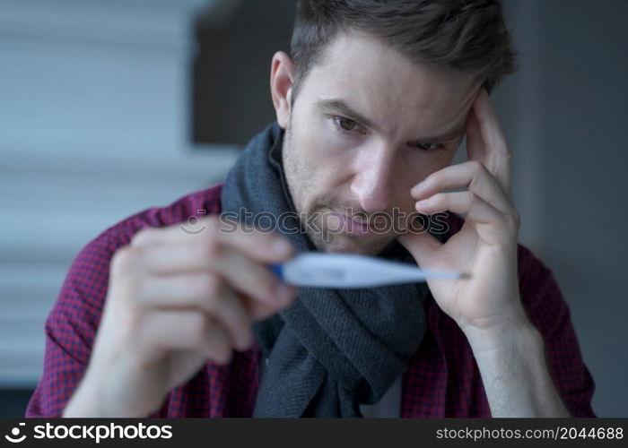 Unhealthy sad young German businessman wearing scarf around neck looking at electronic thermometer, feeling unwell while working in from home, male freelancer suffering of high fever. Unhealthy sad young German businessman wearing scarf around neck looking at electronic thermometer
