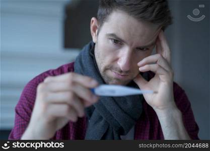 Unhealthy sad young German businessman wearing scarf around neck looking at electronic thermometer, feeling unwell while working in from home, male freelancer suffering of high fever. Unhealthy sad young German businessman wearing scarf around neck looking at electronic thermometer