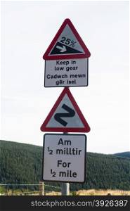 United Kingdom triangular road warning signs in Wales, plates in English and welsh. Steep downhill at 25% and double bend for half a mile.
