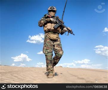 United States marine rider in helmet, camouflage uniform and hidden behind mask and glasses face, carrying satellite communication system on back, walking in desert area with service rifle in hands. Armed marine corps rider patrolling in desert area