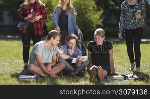 University students studying with tablet pc on university campus while sitting on green grass in park. Handsome male college friends working with touchpad and communicating. Teenage female friends joining classmates, embracing and greeting the boys.