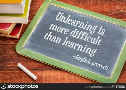 Unlearning is more difficult than learning - English proverb on a slate blackboard with a white chalk and a stack of books against rustic wooden table