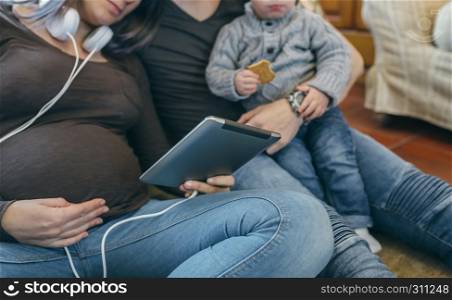 Unrecognizable family with small child and pregnant mother looking at the tablet sitting on the carpet. Family with child and pregnant mother looking tablet