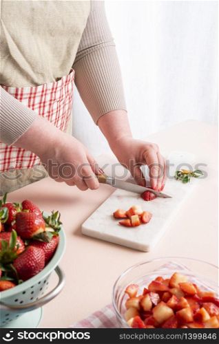 Unrecognizable housewife cutting in small pieces the strawberries. The process of making strawberry jam at home with organic fruits. Woman preparing the fruits for jam