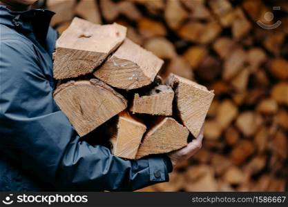 Unrecognizable man carries heap of wood for making fire dressed in jacket. Faceless male carries firewood into house
