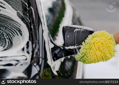 Unrecognizable man washes black car with cloth or rag mitten, uses special detergent. Cleaning dirty automobile. Carwash business concept.
