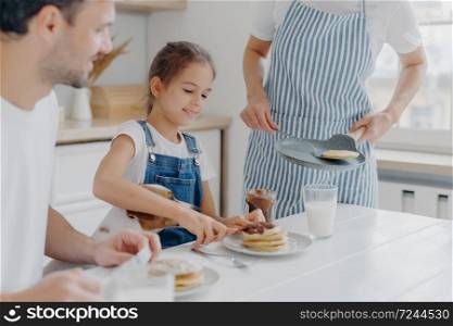 Unrecognizable mommy brings delicious pancakes to table, prepares breakfast for family. Cheerful girl adds melted chocolate to dessert, enjoys time with favourite pet and parents. Breakfast time