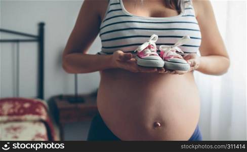 Unrecognizable pregnant woman showing baby sneakers in bedroom. Pregnant showing baby sneakers