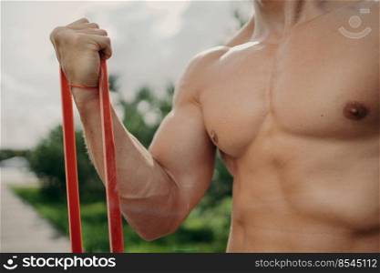 Unrecognizable strong man has workout with resistance band, gets stronger every day, shows well build body, poses outside. Sport, bodybuilding and healthy lifestyle concept. Faceless athlete