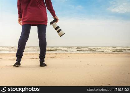 Unrecognizable woman having leisure time walking on beach during autumnal weather holding professional photography camera.. Woman walking on beach with camera