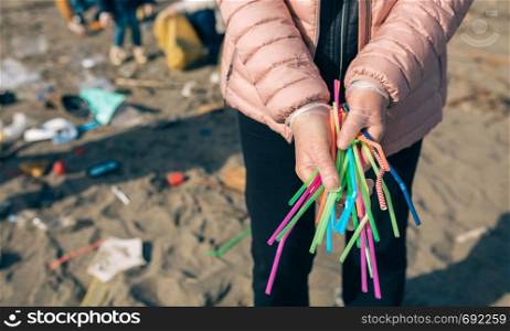 Unrecognizable woman showing handful of straws collected on the beach with group of volunteers working in the background. Selective focus in straws in foreground. Woman showing handful of straws collected on the beach
