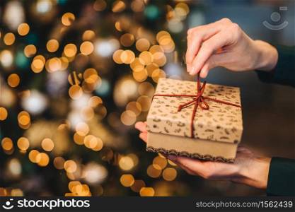 Unrecognizable woman wrappes gift box over Christmas tree with shining lights and garlands. Decorated present box in woman`a hands. Holidays and celebration concept.