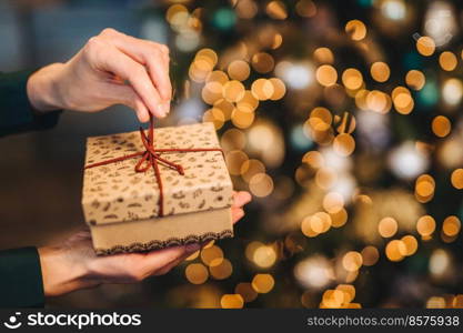 Unrecognizable woman wrappes gift box over Christmas tree with shining lights and garlands. Decorated present box in woman a hands. Holidays and celebration concept.