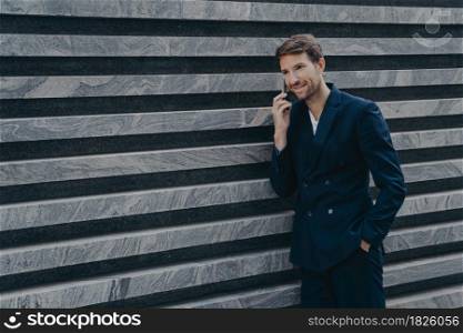 Unshaven male corporate worker dressed in formal black suit keeps hand in pocket has telephone conversation stands near marble wall outdoor. Handsome CEO communicates with professional partner. Male corporate worker dressed in formal black suit keeps hand in pocket has telephone conversation