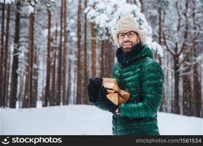 Unshaven smiling male holds pile of firewood, going to make fire to warm himself in frosty weather, spend winter day in forest, has picnic with friends outdoor. Fashionable unshaven man chop firewood