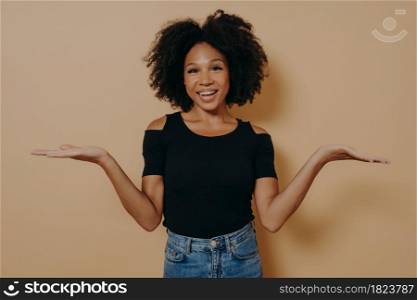 Unsure african female model in with curly hair shrugging shoulders with uncertainty with clueless and confused look while posing alone isolated over beige background, dressed in casual outfit. Unsure african female model shrugging shoulders with uncertainty with clueless and confused look