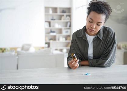 Unwell african american girl is holding pills flacon. Sick teenage girl is taking vitamins or painkillers. Young woman is upset and worried. Stressed patient. Pharmaceutical treatment concept.. Unwell stressed african american girl is holding pills flacon. Pharmaceutical treatment concept.