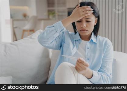 Unwell sleepy woman is examining temperature and touches head. European girl is sitting on couch at home and holding thermometer. Fatigue from virus infection, influenza. Sick leave concept.. Unwell sleepy woman is examining temperature and touches head. Fatigue from virus infection.