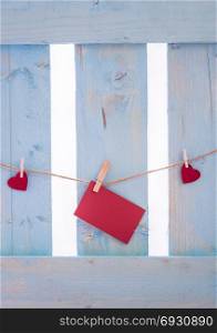 Unwritten red paper note surrounded by red hearts, tied to a linen string with wooden clips, on a blue fence.