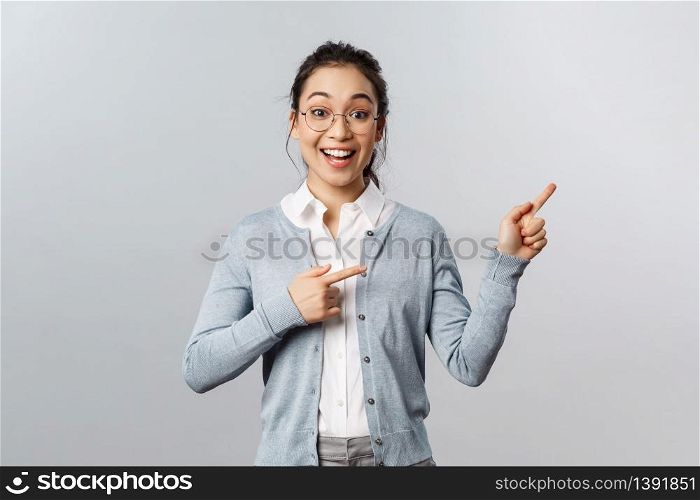 Upbeat excited mixed-race girl in glasses, asian woman teacher pointing fingers right, smiling broadly, found way to stay in touch with students during lockdown quarantine, standing grey background.. Upbeat excited mixed-race girl in glasses, asian woman teacher pointing fingers right, smiling broadly, found way to stay in touch with students during lockdown quarantine, standing grey background