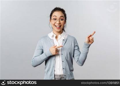 Upbeat excited mixed-race girl in glasses, asian woman teacher pointing fingers right, smiling broadly, found way to stay in touch with students during lockdown quarantine, standing grey background.. Upbeat excited mixed-race girl in glasses, asian woman teacher pointing fingers right, smiling broadly, found way to stay in touch with students during lockdown quarantine, standing grey background