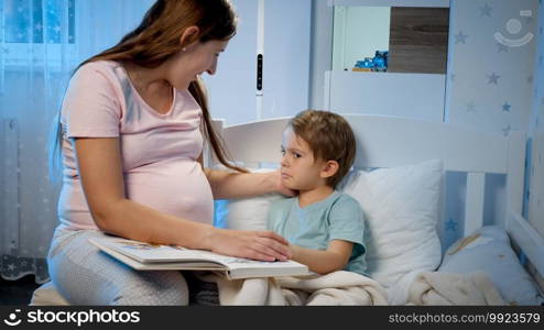 Upset and crying toddler boy lying in bed and hugging his pregnant mother sitting next to his bed at night.. Upset and crying toddler boy lying in bed and hugging his pregnant mother sitting next to his bed at night