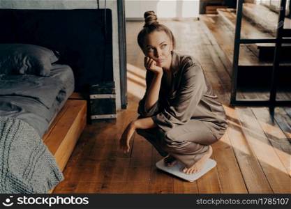 Upset and unmotivated woman on electronic floor scale in bedroom wondering why cannot lose weight despite her new diet, looking aside with sade face expression. Weight Loss and healthy lifestyle. Upset young attractive woman using scales at home and wondering why cannot lose weight
