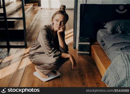 Upset and unmotivated woman on electronic floor scale in bedroom wondering why cannot lose weight despite her new diet, looking aside with sade face expression. Weight Loss and healthy lifestyle. Upset young attractive woman using scales at home and wondering why cannot lose weight