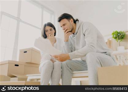 Upset couple stressed by bad news, receive high taxes, looks attentively at papers, read paper letter, have to pay much for flat, move in other apartment, pack belongings in cardboard containers