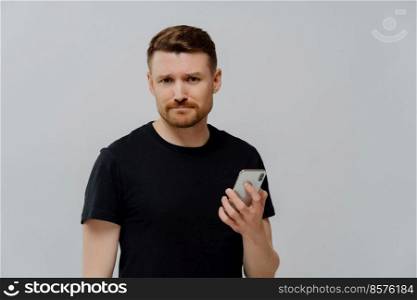Upset dissatisfied bearded young man holds modern smartphone waits for call frowns face dressed in black casual t shirt isolated over blue background being worried about recent terrifying news