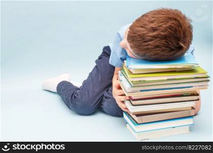 Upset schoolboy sitting with pile of school books. boy sleeping on a stack of textbooks isolated on a blue background. Upset schoolboy sitting with pile of school books. boy sleeping on a stack of textbooks
