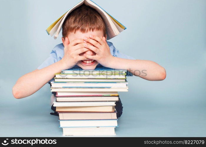 Upset schoolboy sitting with pile of school books and covers his face with hands isolated on a blue background. schoolboy sitting with pile of school books and covers his face with hands isolated on a blue background