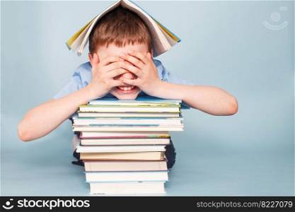 Upset schoolboy sitting with pile of school books and covers his face with hands isolated on a blue background. schoolboy sitting with pile of school books and covers his face with hands isolated on a blue background