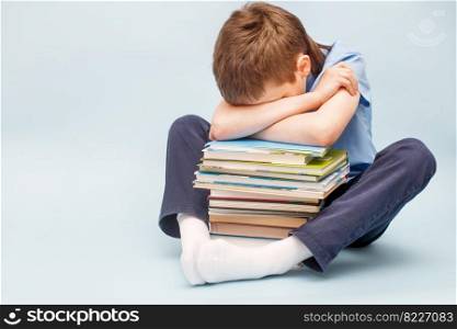 Upset schoolboy sitting with pile of school books and covers his face with hands. boy sleeping on a stack of textbooks isolated on a blue background. Upset schoolboy sitting with pile of school books and covers his face with hands. boy sleeping on a stack of textbooks