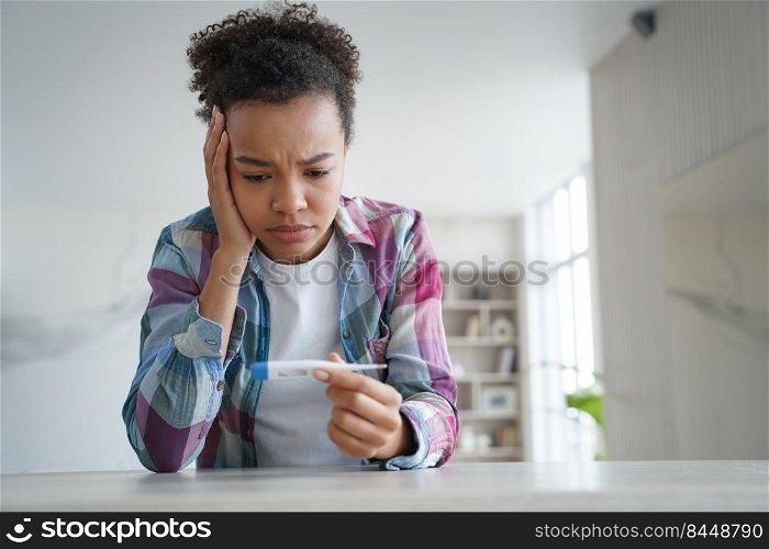 Upset unhealthy young african american woman looks at electronic thermometer. Biracial teen girl dissapointed with high body temperature covid symptom, suffer from fever, caught cold before trip.. Sad african american girl looks at thermometer, has high body temperature, fever, covid symptom