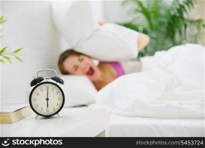 Upset woman closing ears by pillow to avoid hearing alarm clock