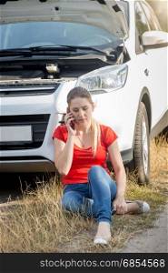 Upset woman sitting on ground and leaning on broken car